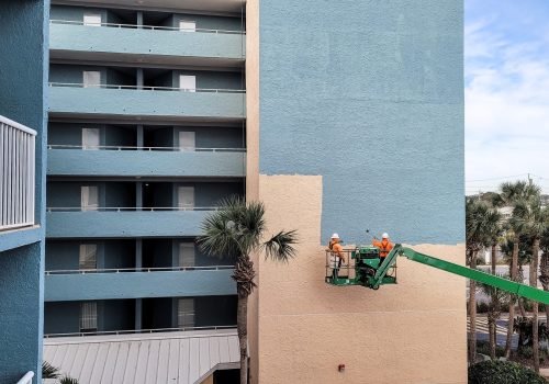 Crane in heavy machinery lifts construction workers up side of building to paint new blue color.